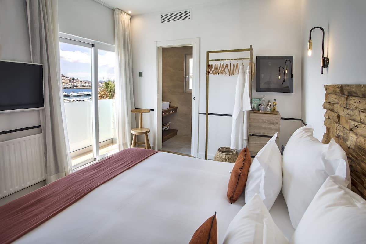 A hotel room with a large bed, a TV, a view of the sea, a wooden wall, and a bathroom with a towel rack.