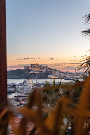 A coastal town with a prominent castle-like structure on a hill, boats docked at a marina, and palm trees in the foreground.