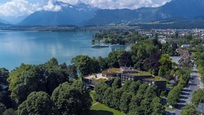 A lakeside town with a prominent building featuring a green roof, surrounded by lush greenery and a view of a calm lake with boats. - Hotel Seepark Thun (Thun)