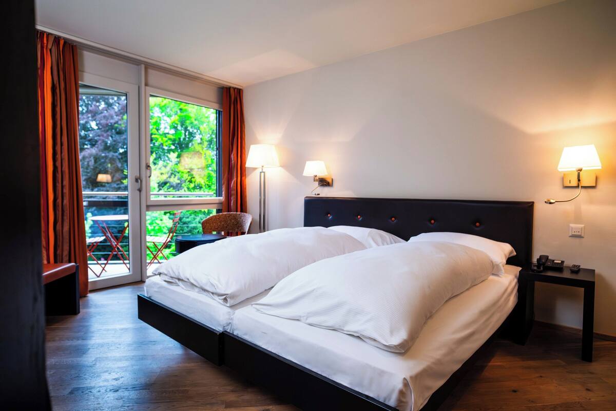 A bedroom with a bed, two bedside lamps, a nightstand, a window with curtains, and a view of greenery outside.