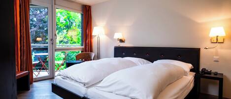 Ein Schlafzimmer mit einem Bett, zwei Nachttischlampen, einem Nachttisch, einem Fenster mit Vorhängen und Blick auf Grünfläche.