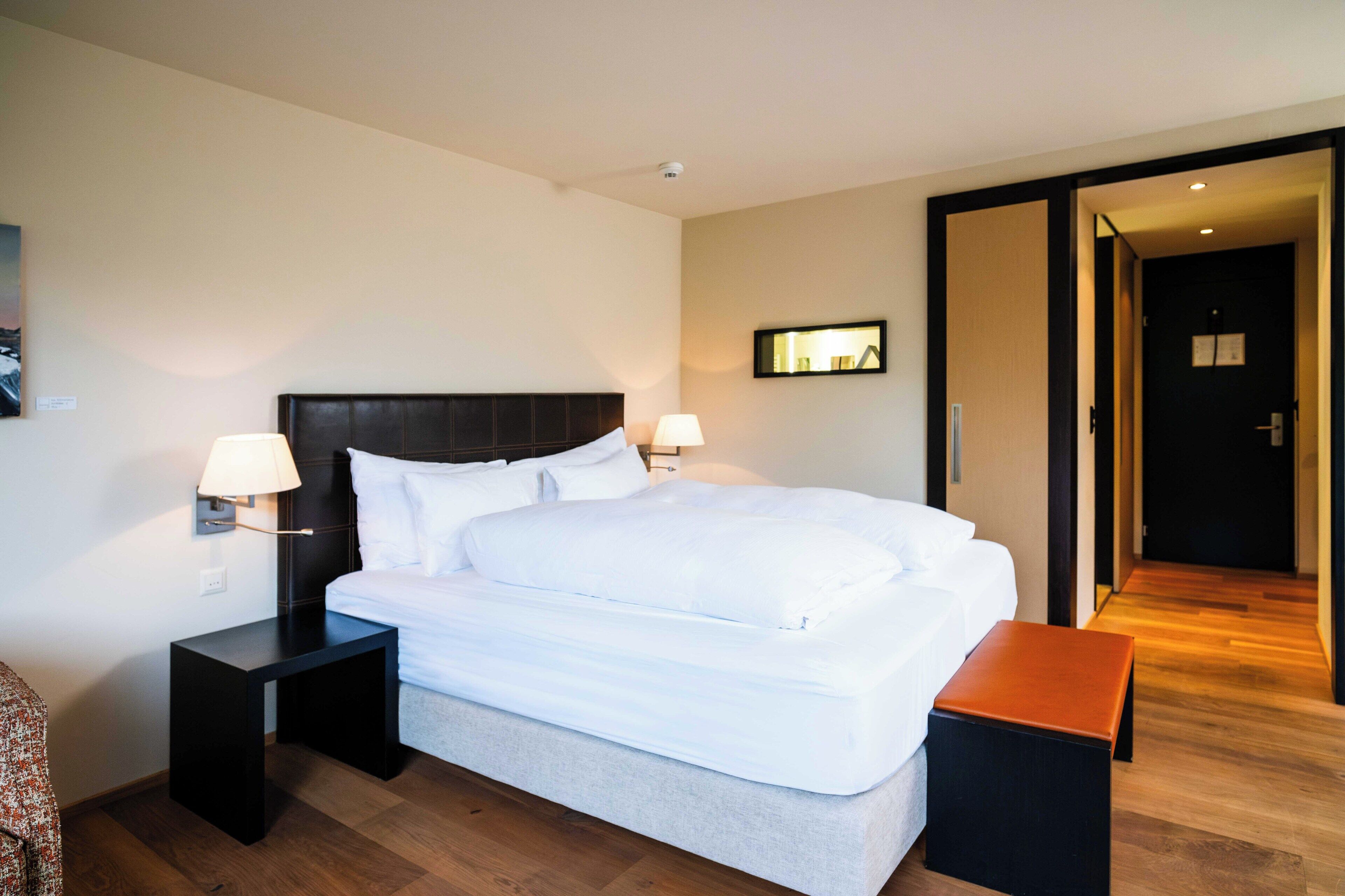 A hotel room with a large bed, two bedside tables, a bench, a wall-mounted lamp, and a door leading to another room.