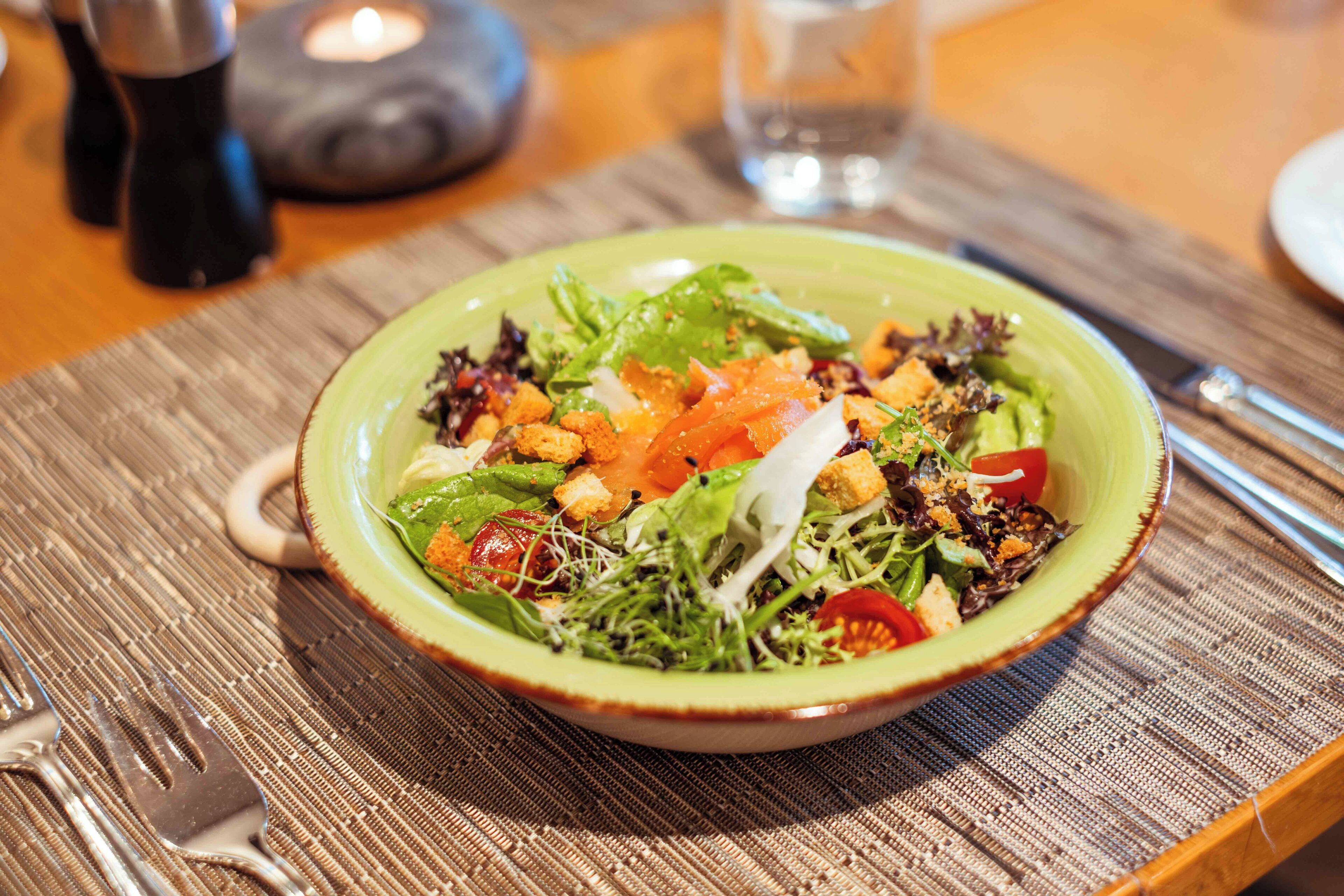 A green bowl of salad on a wooden table with a fork and a glass of water.