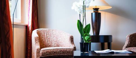 A room with a patterned armchair, a small table with a lamp and a potted orchid, and a window with curtains.