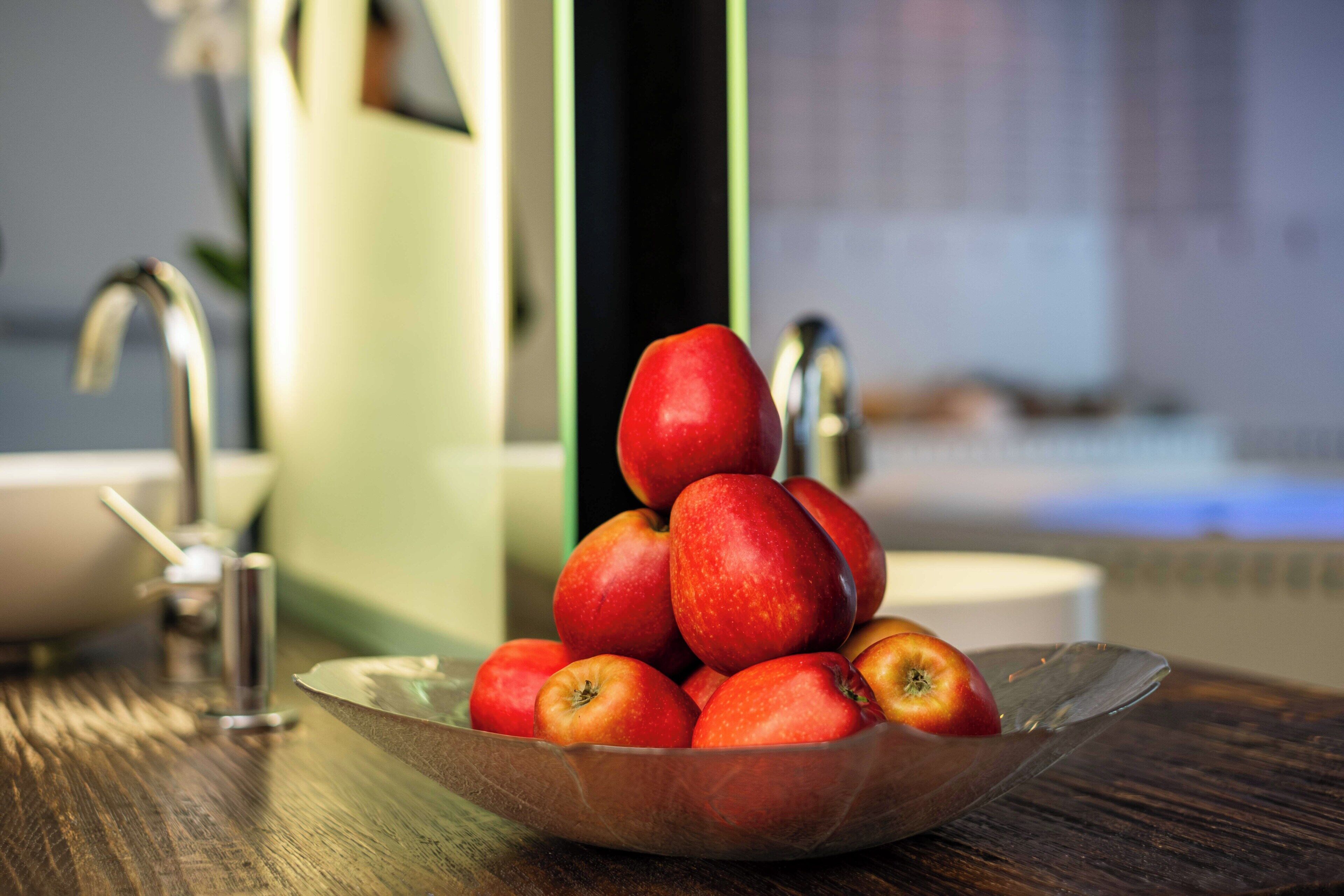 A bowl of apples on a wooden table in a bathroom with a sink and mirror.