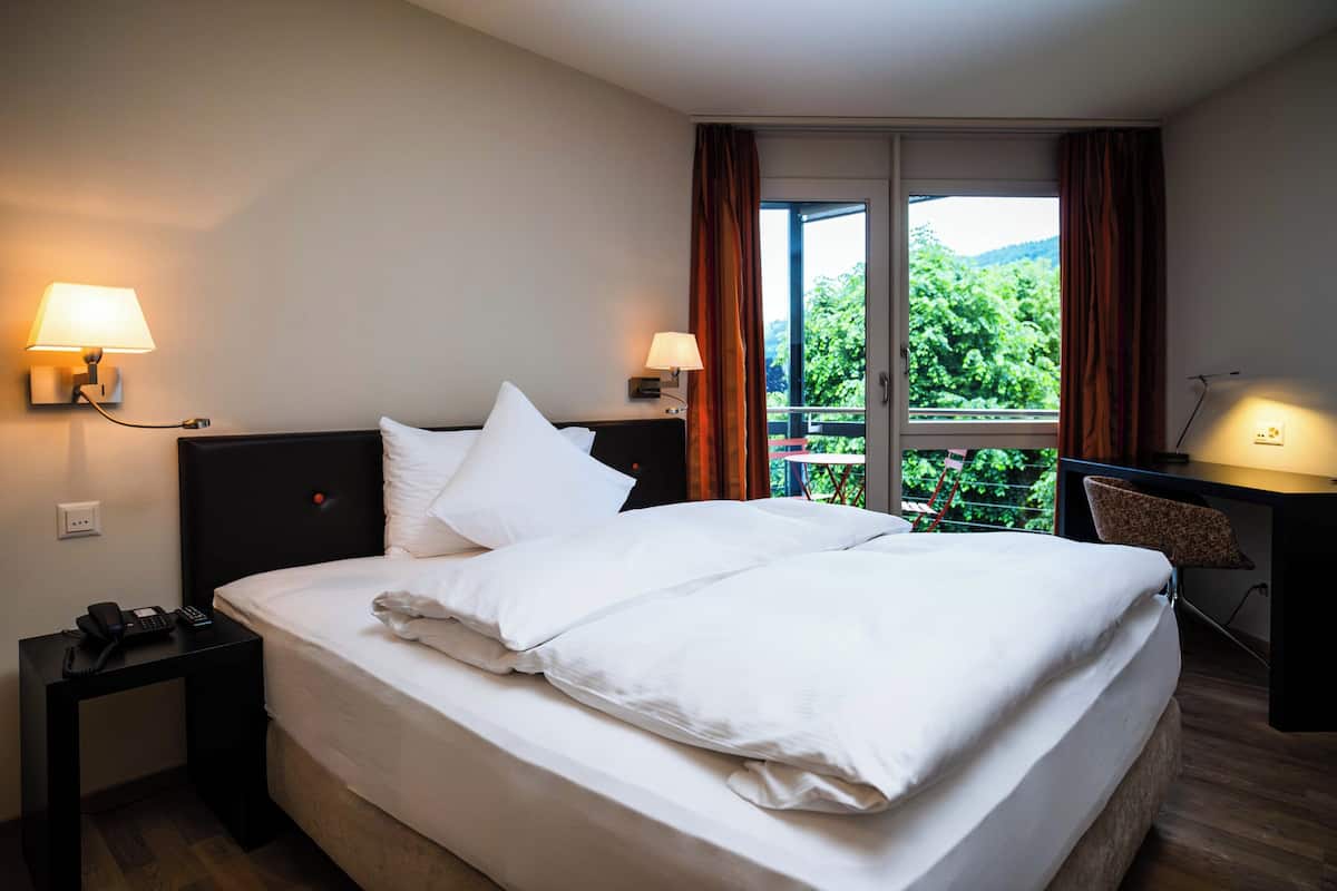 A hotel room with a large bed, two bedside lamps, a desk with a chair, and a window with curtains.