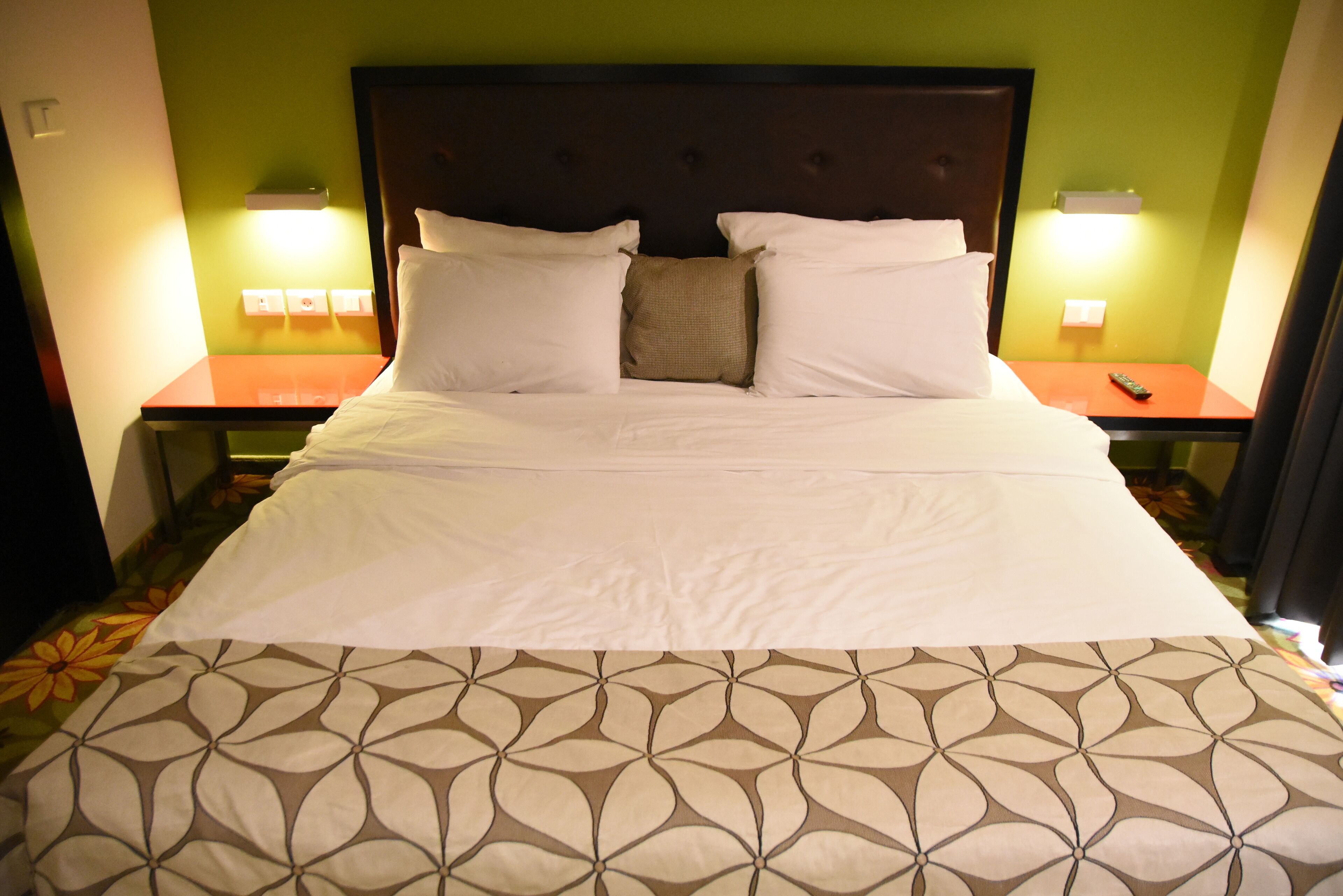 Una cama bien hecha con ropa de cama blanca y estampada, dos mesitas de noche con lámparas y una pared verde.
