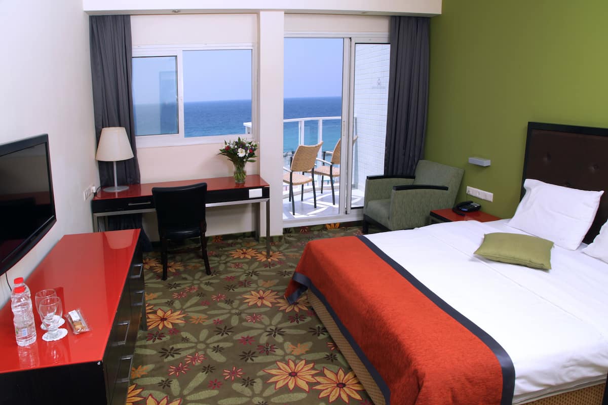 A hotel room with a bed, desk, and chairs. There is a view of the ocean through the window.