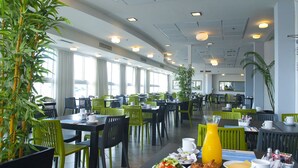 A spacious dining area with multiple tables and chairs, large windows, and indoor plants. - Madison Hotel Nahariya (Nahariya)