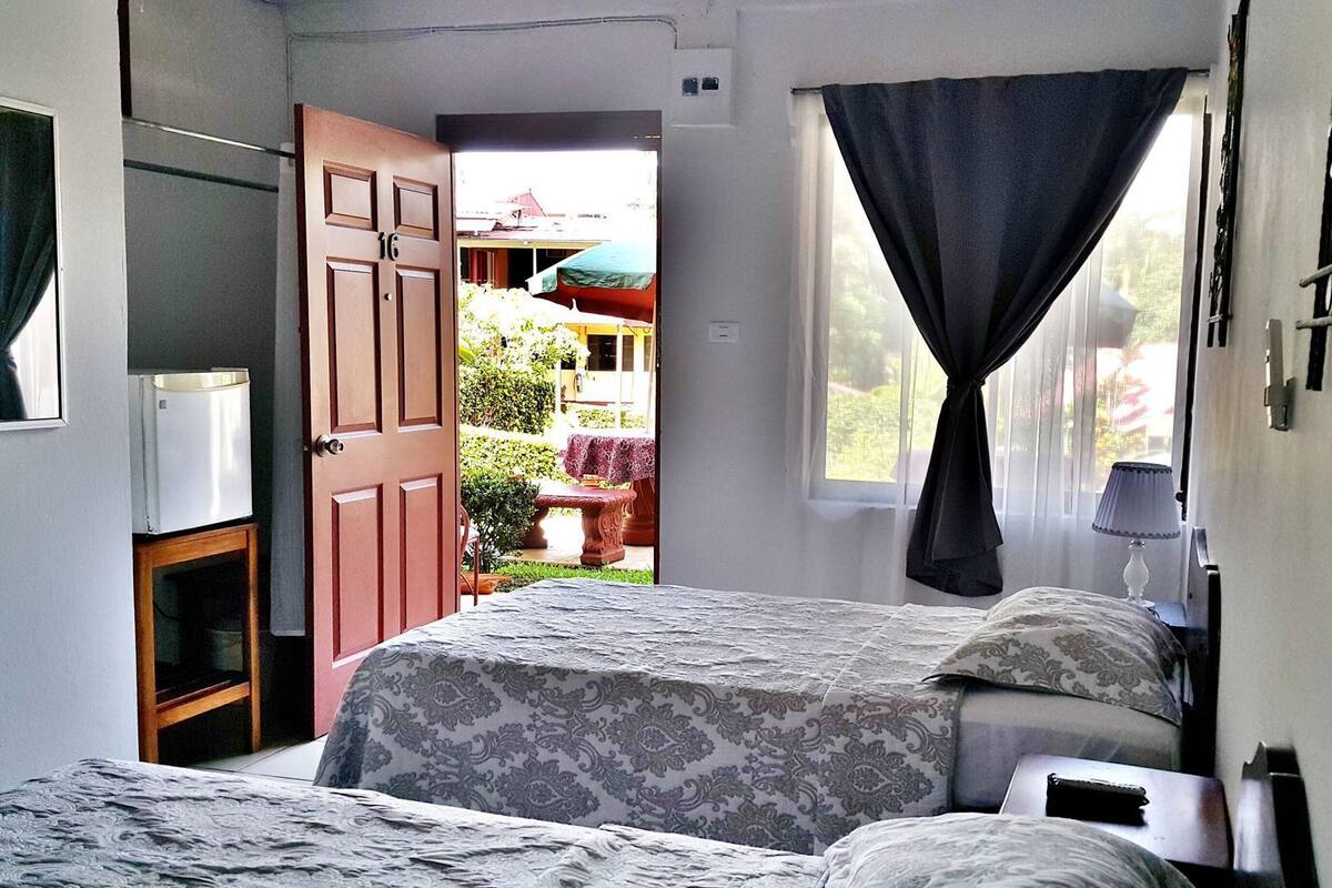 Quarto com duas camas, uma janela com cortinas, uma porta para o exterior e uma pequena mesa com abajur.