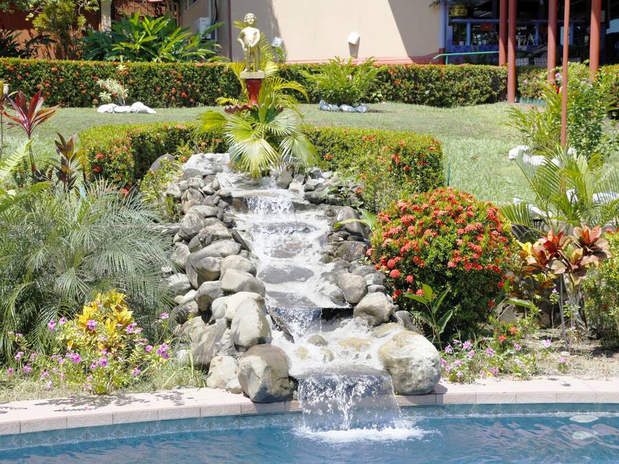 Una piscina con cascada rodeada de plantas tropicales y una estatua.