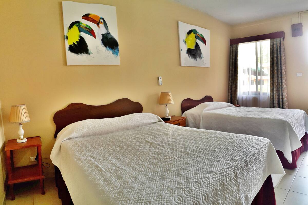 Quarto de hotel com duas camas, uma mesa de cabeceira com abajur e dois quadros de araras nas paredes.