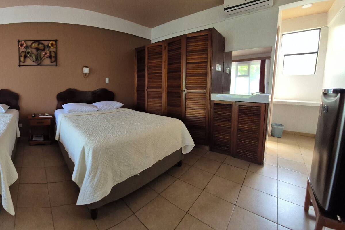 Quarto com duas camas, uma porta com identificação, uma luminária na parede e uma janela com cortinas.