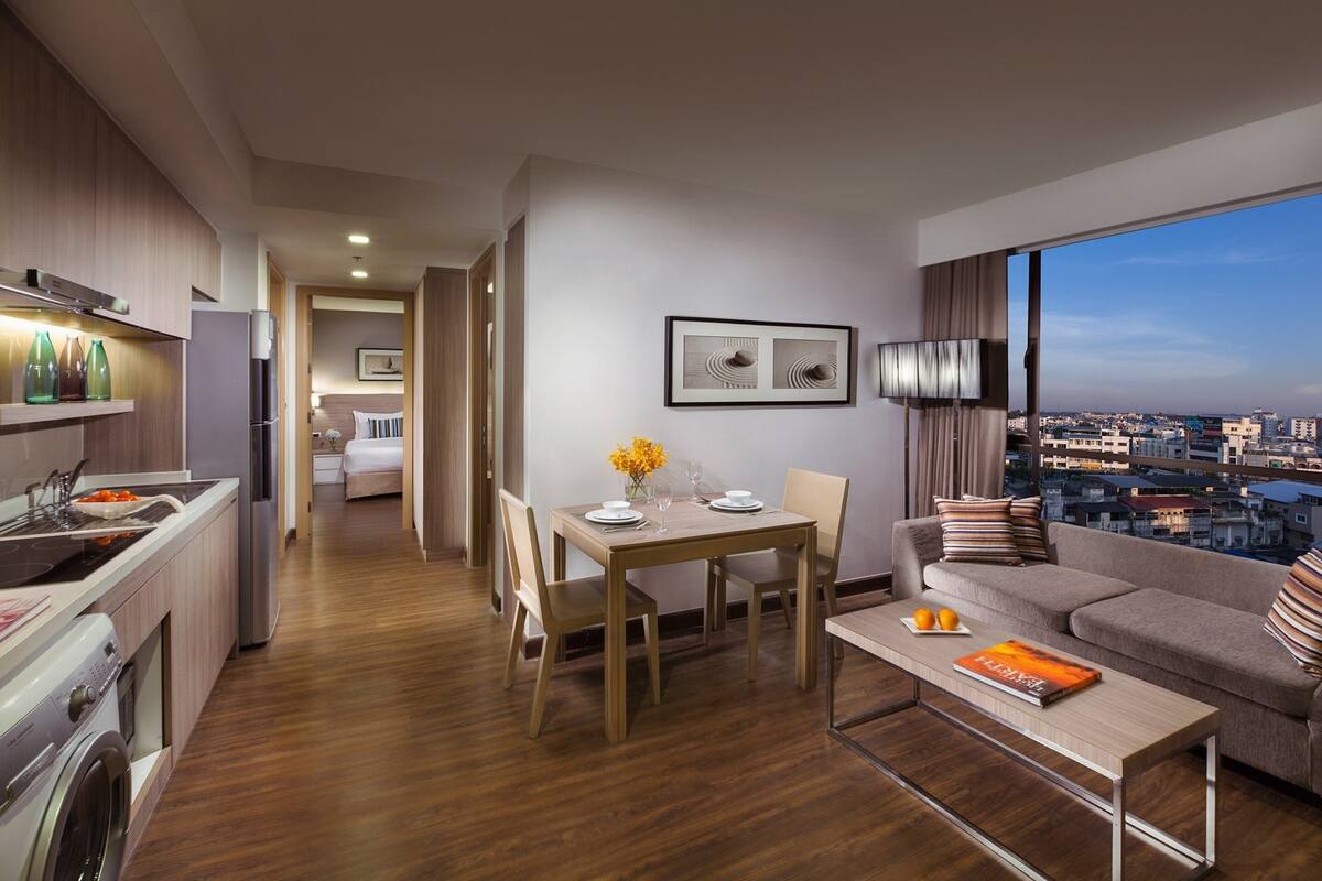 A modern hotel room with a dining area, kitchenette, and a sofa with a city view.