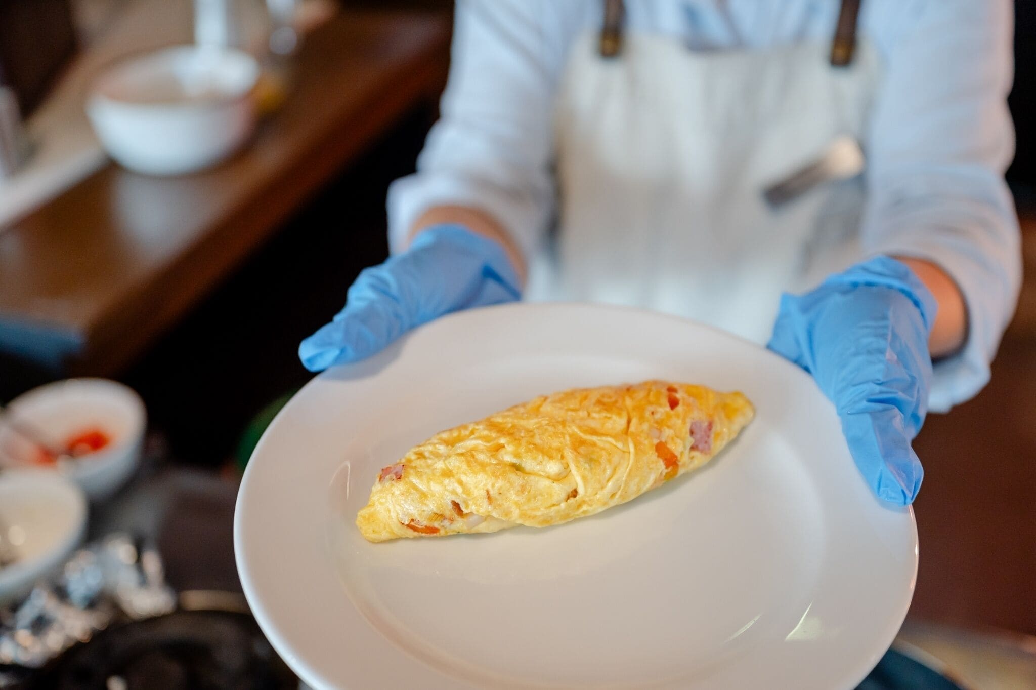 A person in a white coat and blue gloves holding a plate with an omelette.