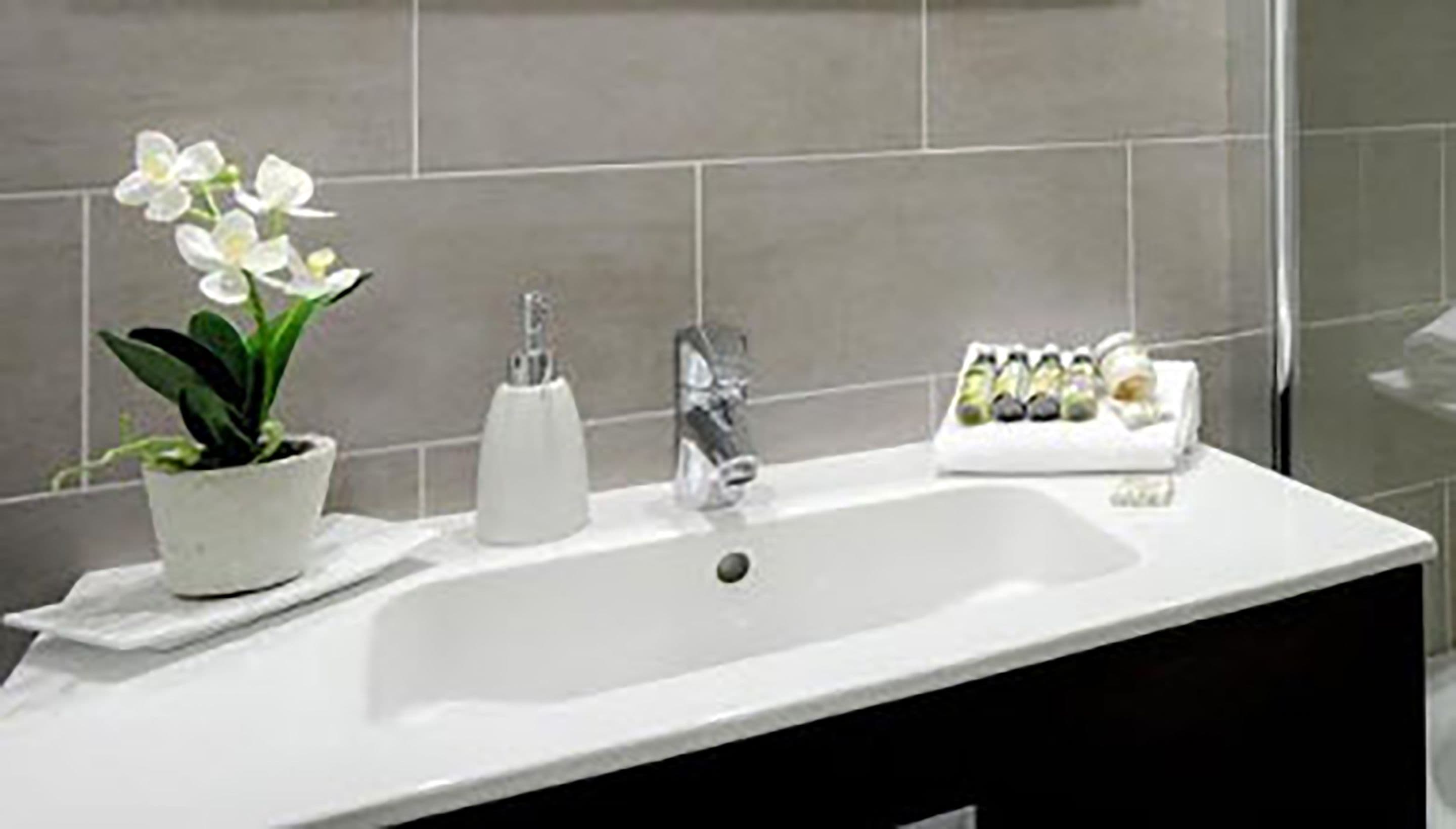 Une salle de bain avec un lavabo blanc, une orchidée en pot, un distributeur de savon et un plateau contenant des articles de toilette.