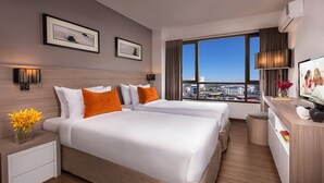 Ein Hotelzimmer mit zwei Betten, einem Schreibtisch, einem Fernseher und einem großen Fenster mit Blick auf die Stadt.