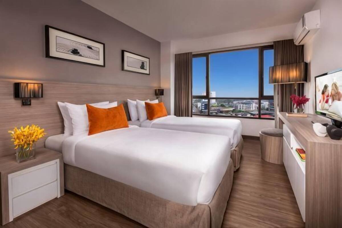A hotel room with two beds, a desk, a chair, a TV, and a large window with a city view.