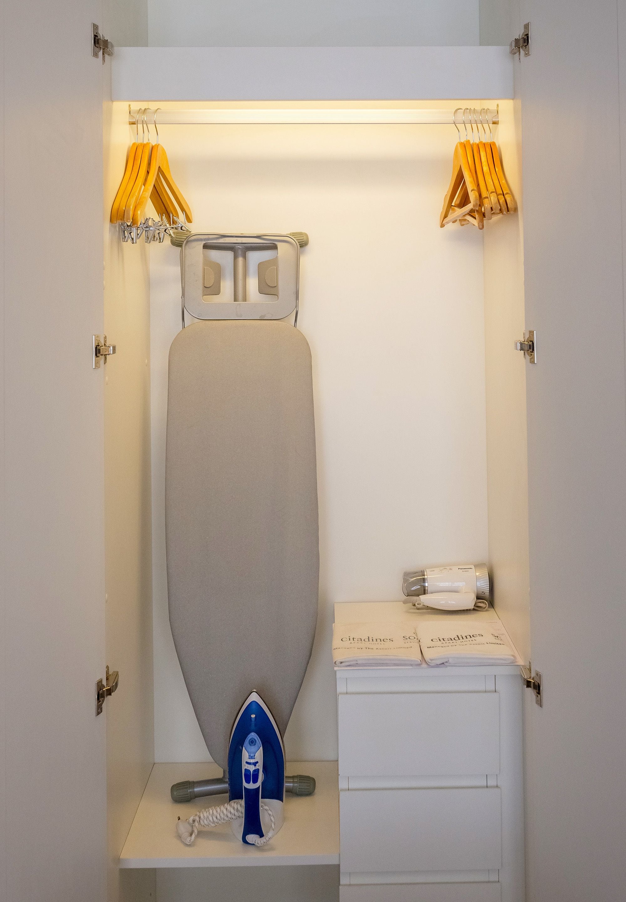 Une pièce compacte et bien éclairée, dotée d’un espace intégré pour le repassage, d’une petite table avec un téléphone et d’une étagère où sont suspendus des vêtements.