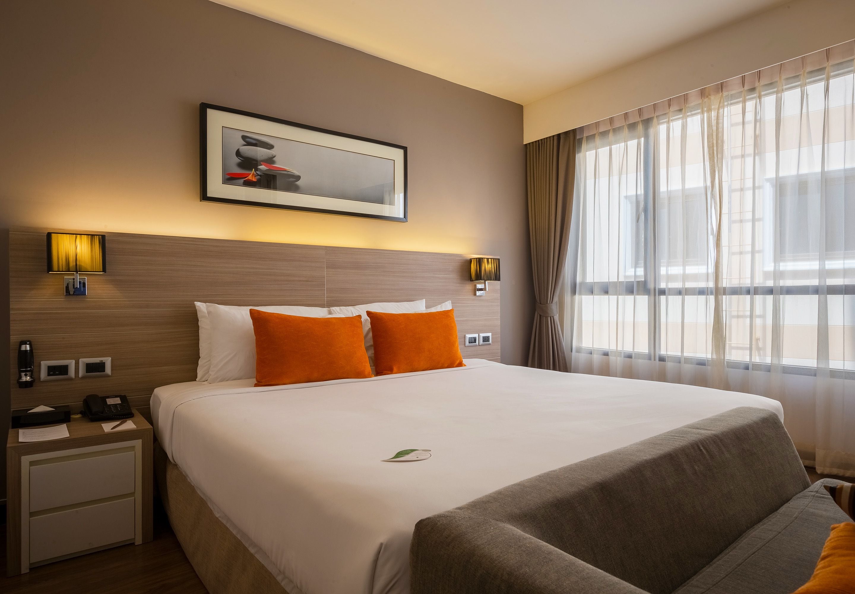 Una habitación de hotel con una cama grande, dos lámparas de noche, una mesita de noche, un cuadro en la pared y una ventana con cortinas translúcidas.