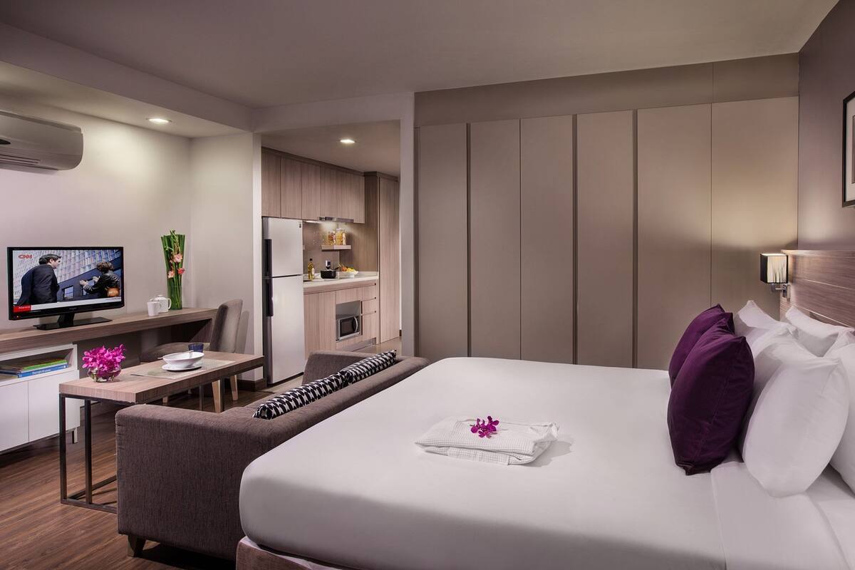 A modern hotel room with a bed, a desk, a television, and a kitchenette.