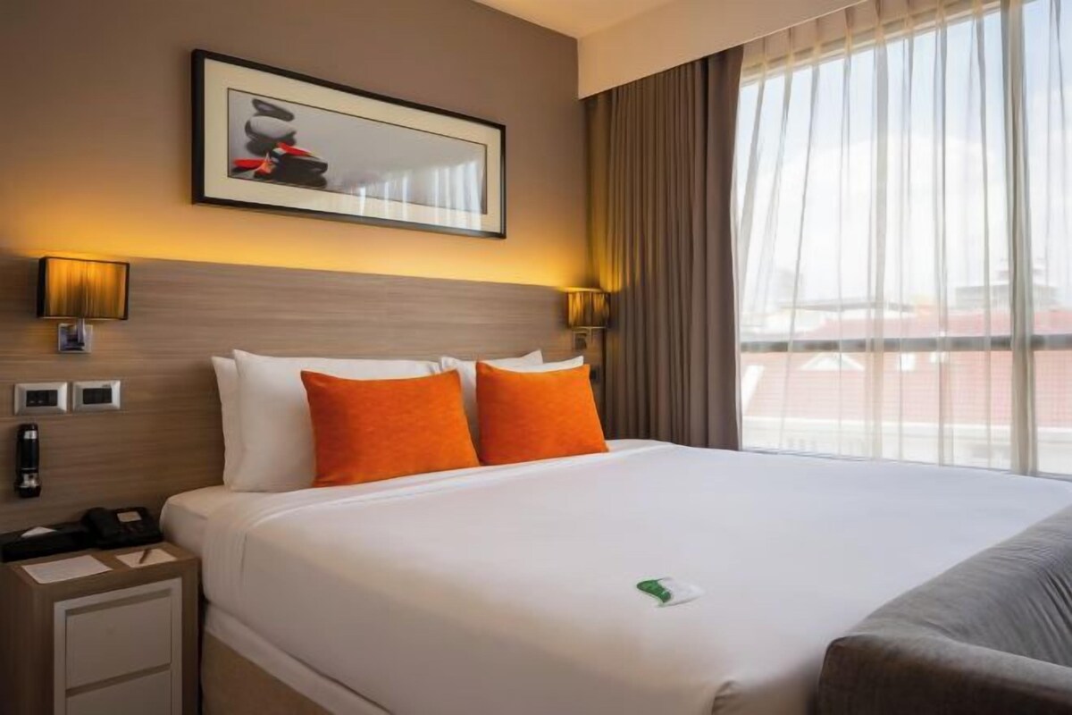 A hotel room with a large bed, two orange pillows, a framed picture on the wall, and a window with curtains.