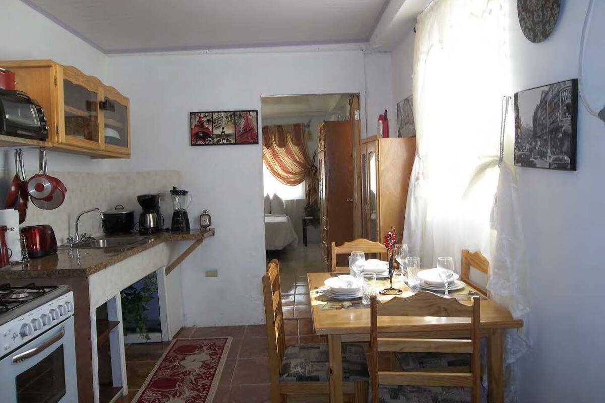 Une cuisine compacte avec des meubles en bois, une cuisinière et divers appareils électroménagers. On aperçoit un coin repas avec une table et des chaises, qui donne sur une chambre à coucher équipée d’un lit et de rideaux.