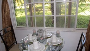 A dining table set with white plates, glasses, and a pitcher, positioned by a window with a view of a grassy area and trees. - Tobago Tranquil Guesthouse (Buccoo)
