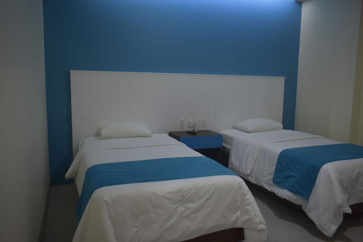Habitació d'hotel amb dos llits, cada un amb roba de llit blanca i detalls blaus, una tauleta de nit amb una làmpada i una paret d'accent blava.
