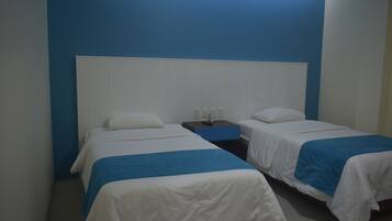 Habitación de hotel con dos camas, cada una con ropa de cama blanca y detalles azules, una mesita de noche con lámpara y una pared de acento azul.