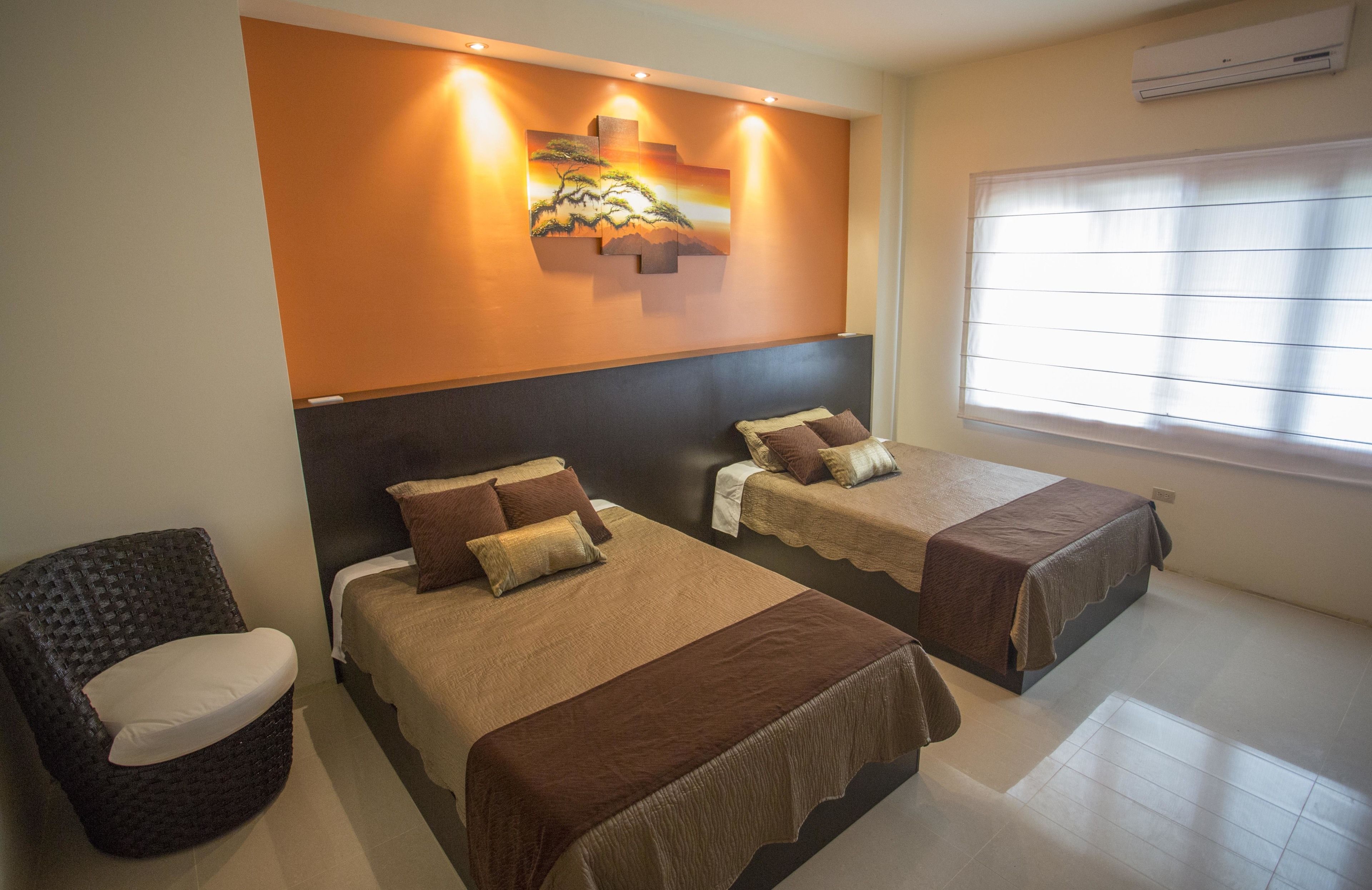 A hotel room with two beds, a chair, a painting on the wall, and a window with blinds.