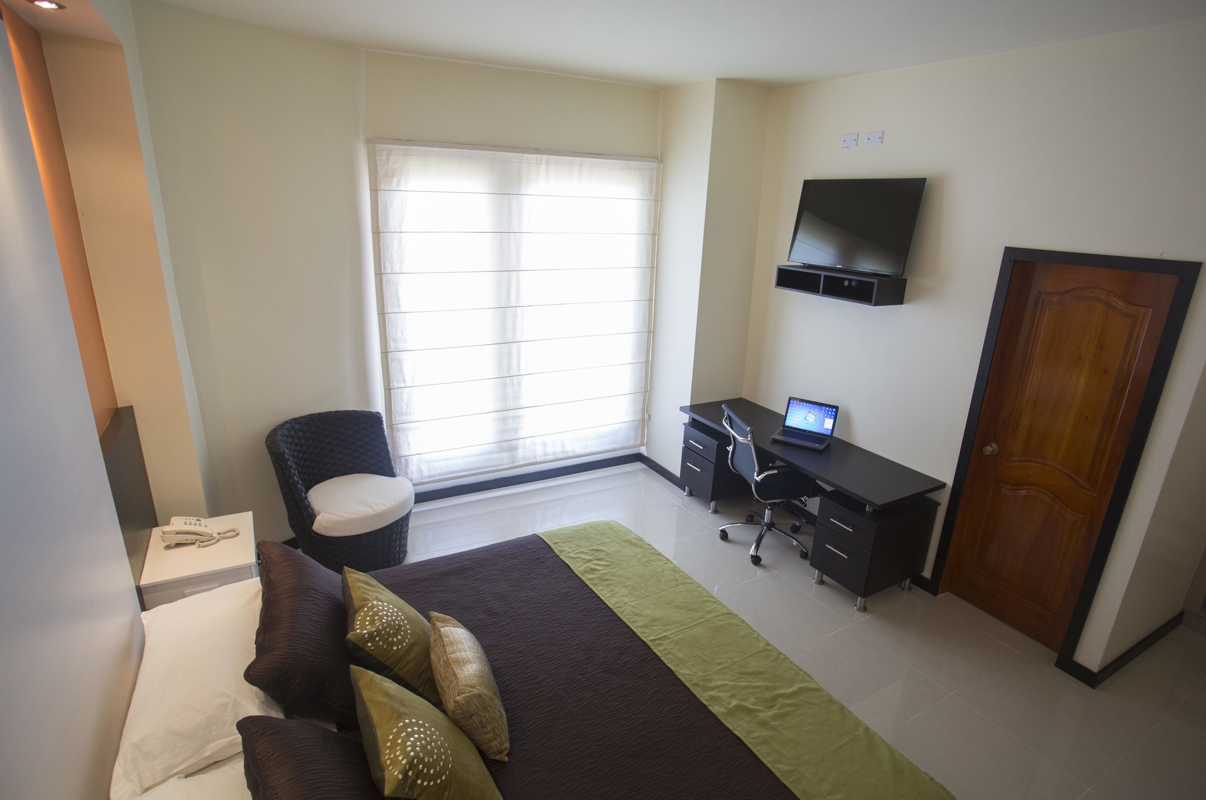 A bedroom with a bed, a desk, a chair, a TV, and a window.