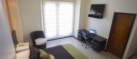 A bedroom with a bed, a desk, a chair, a TV, and a window.