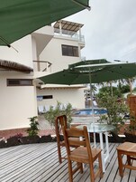 Un área junto a la piscina con sillones de madera, una mesa de vidrio y sombrillas verdes.