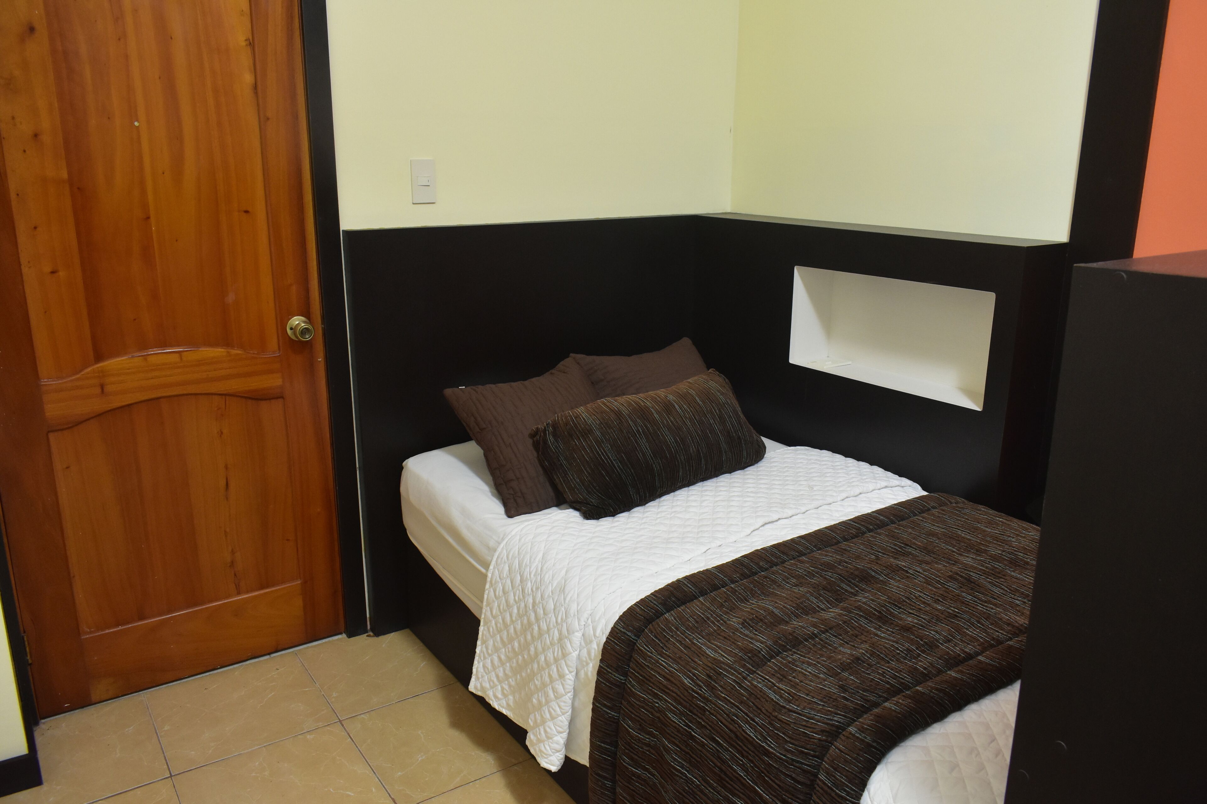 A small, compact bedroom with a wooden door, a bed with a white quilt, and a brown headboard.