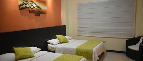 A hotel room with two beds, each with a green blanket and white bedding. There is a painting on the wall and a large window with blinds.