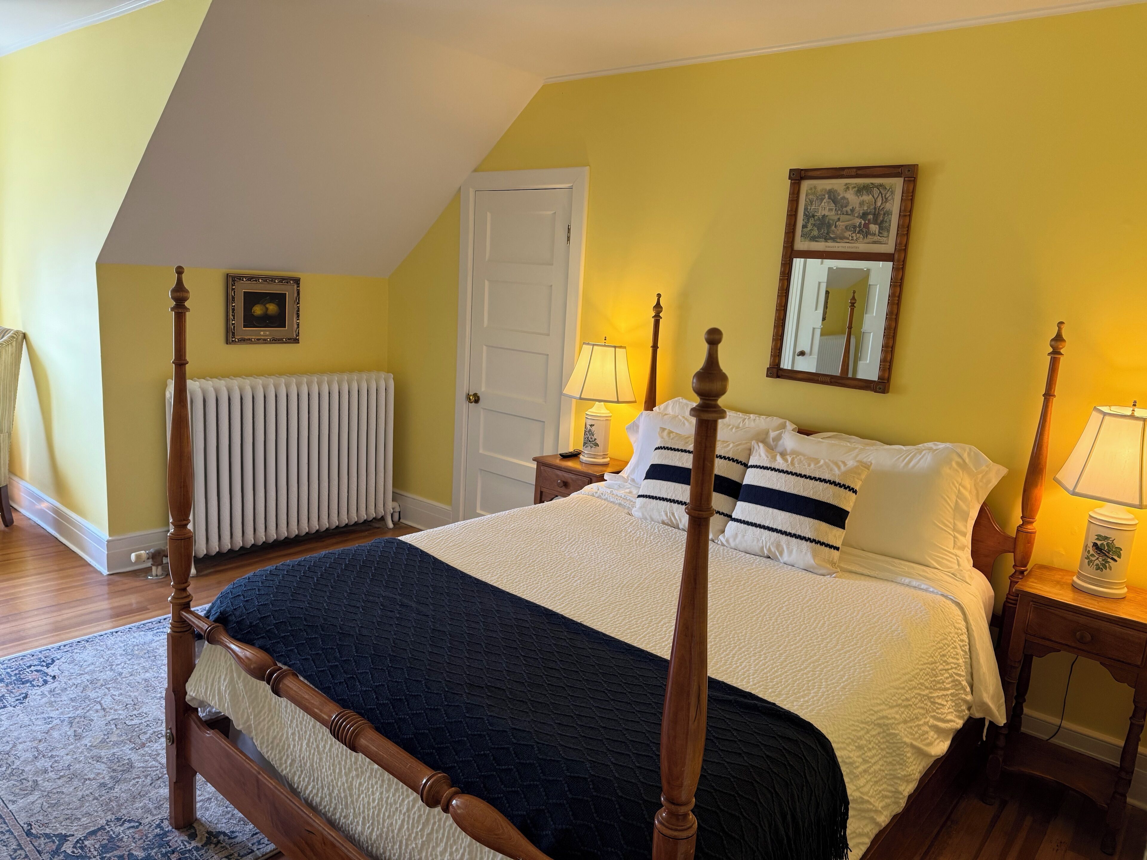 A bedroom with a four-poster bed, a white door, a radiator, a bedside table with a lamp, and a framed picture on the wall.