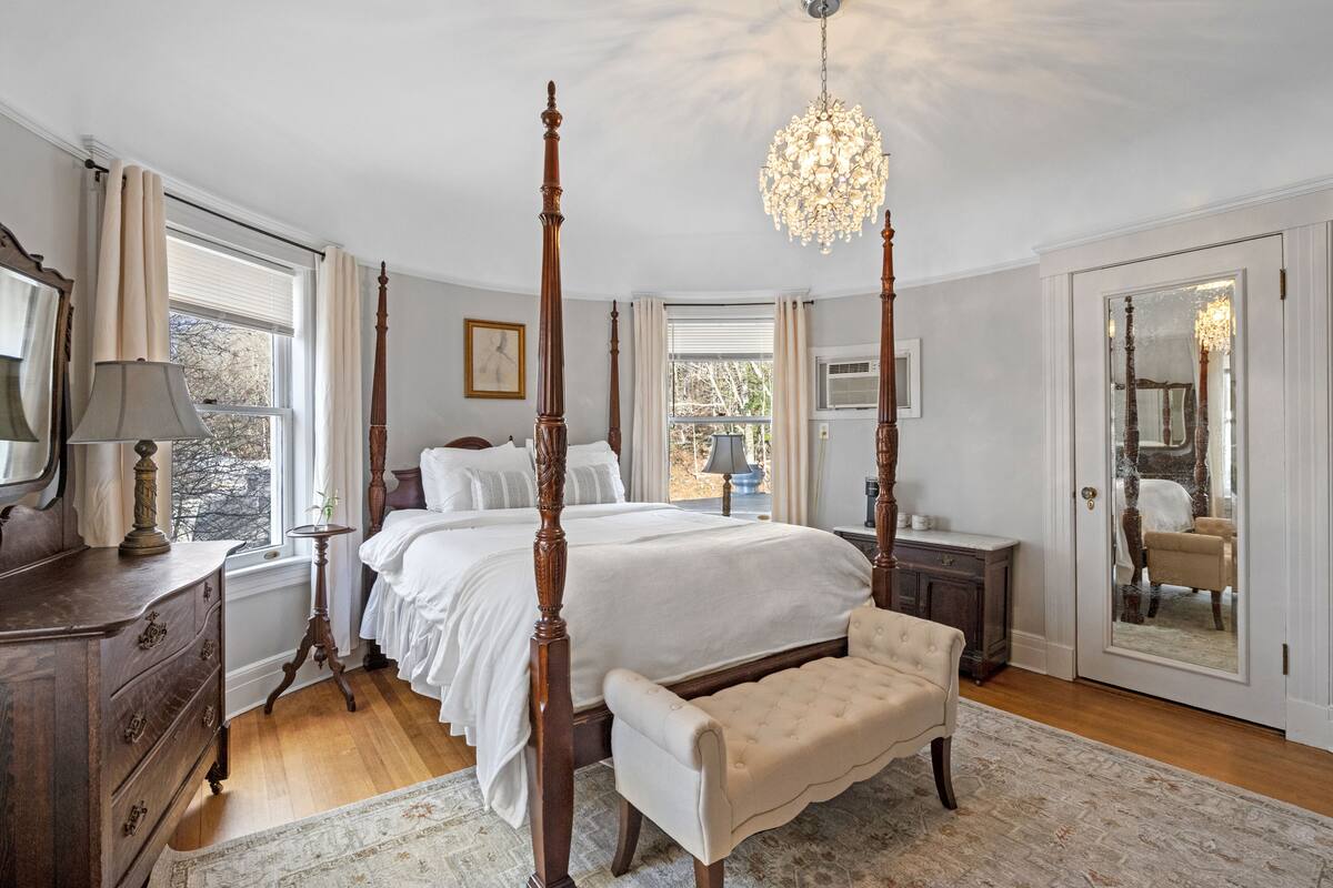A four-poster bed with a chandelier, a wooden dresser, a bench, and a large mirror.