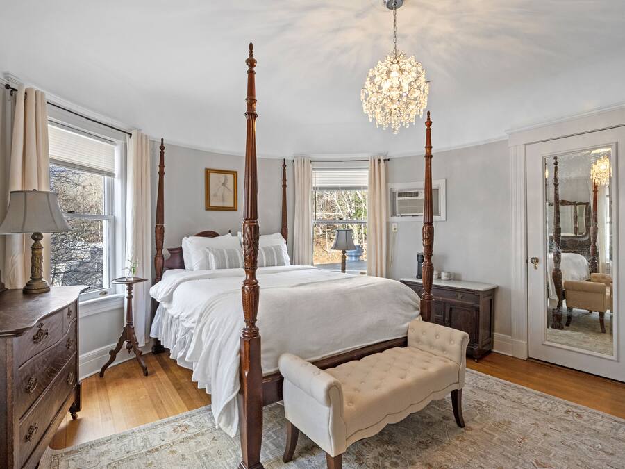 A four-poster bed with a chandelier, a wooden dresser, a bench, and a large mirror.