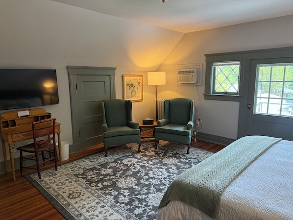 A room with a bed, two armchairs, a wooden desk, a television, an air conditioning unit, and a window with a view of greenery.