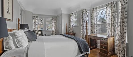 Un dormitorio con una cama grande, un aparador de madera y un escritorio. Hay dos ventanas con cortinas florales.