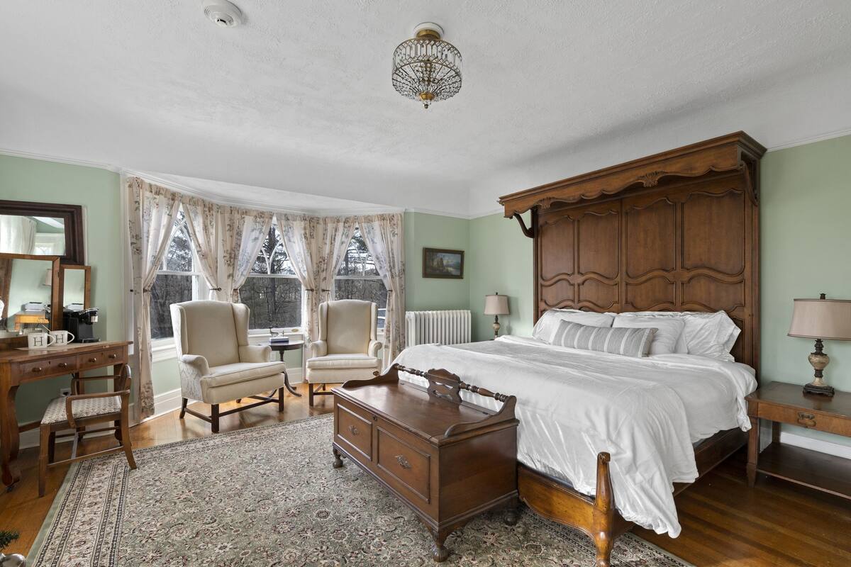 A bedroom with a four-poster bed, a wooden dresser, a desk with a chair, and a large window with curtains.