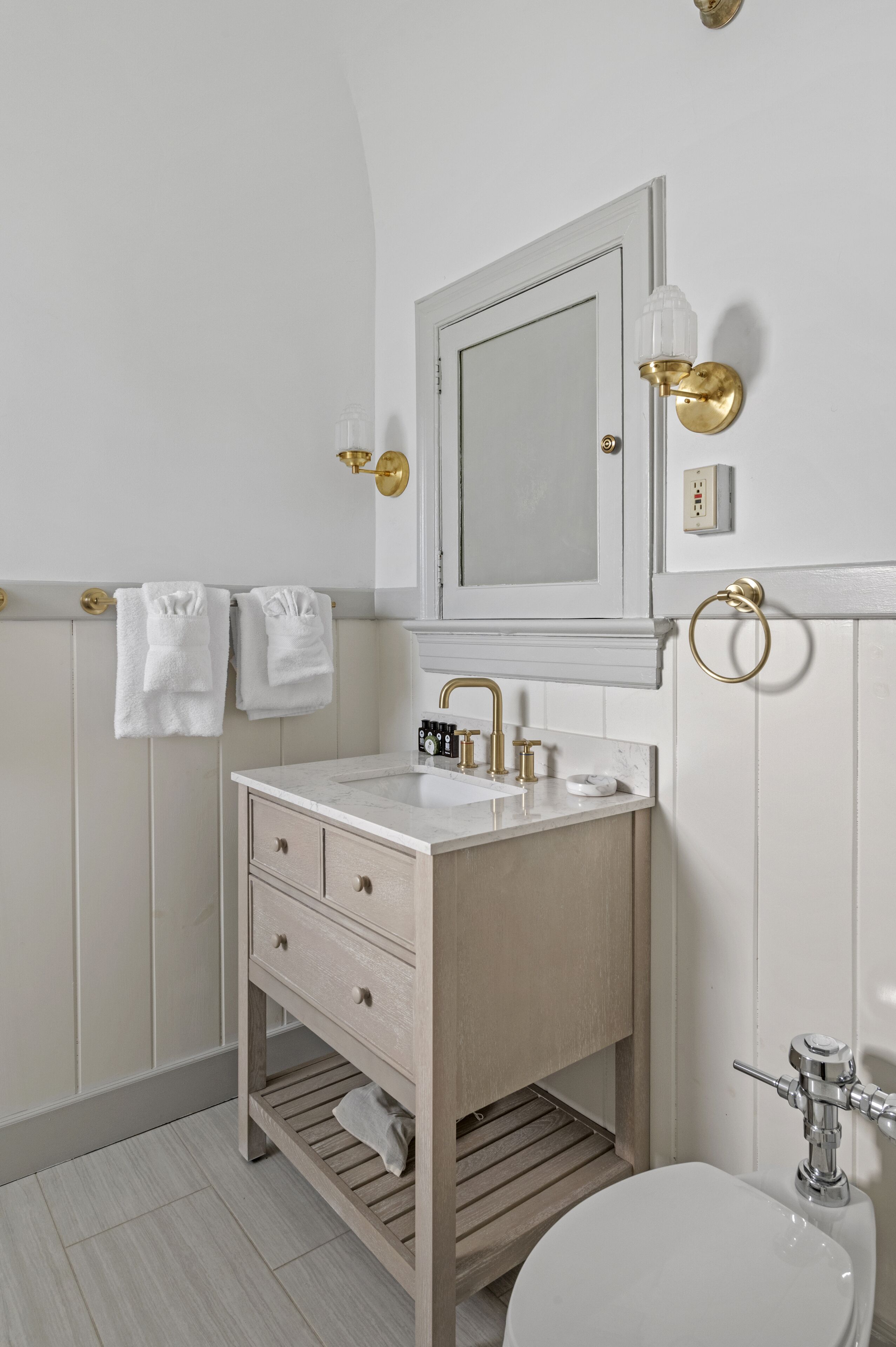 Une salle de bain avec un meuble-lavabo blanc, un miroir, un évier et des toilettes. Des serviettes sont accrochées au mur et un luminaire est fixé au-dessus du miroir.