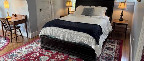 A bedroom with a large bed, two bedside lamps, a desk with a chair, a door, and a window with a view of greenery.