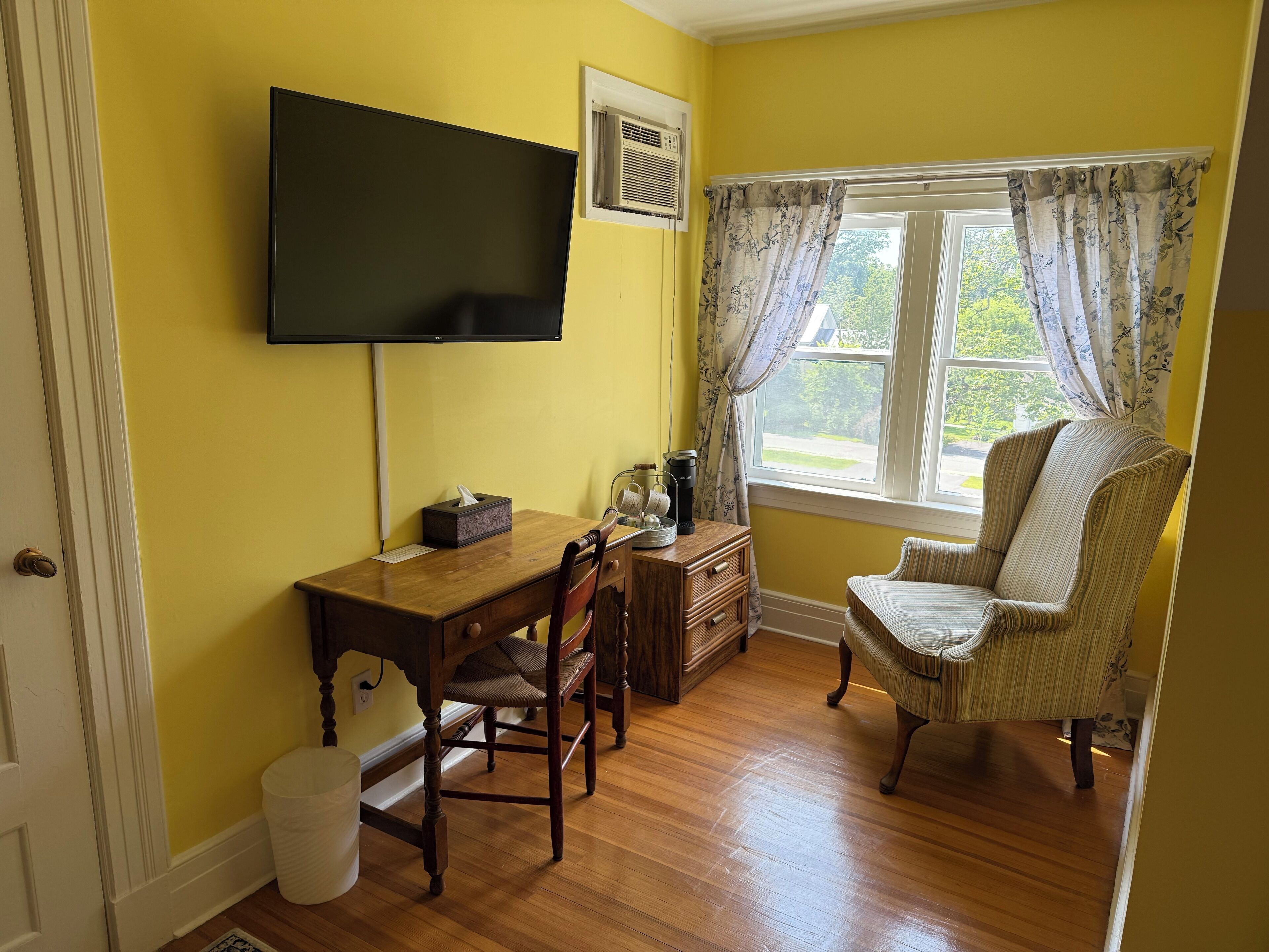 A room with yellow walls, a wooden desk with a chair, a television mounted on the wall, a window with floral curtains, and a striped armchair.