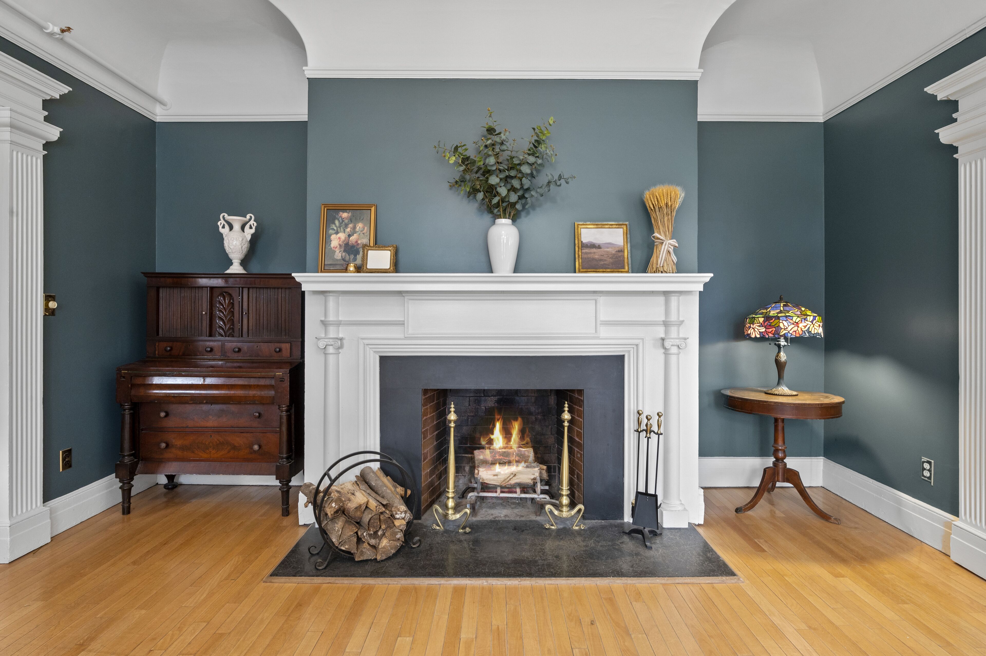 A room with a fireplace, a wooden dresser, a vase with flowers, and a small table with a lamp.