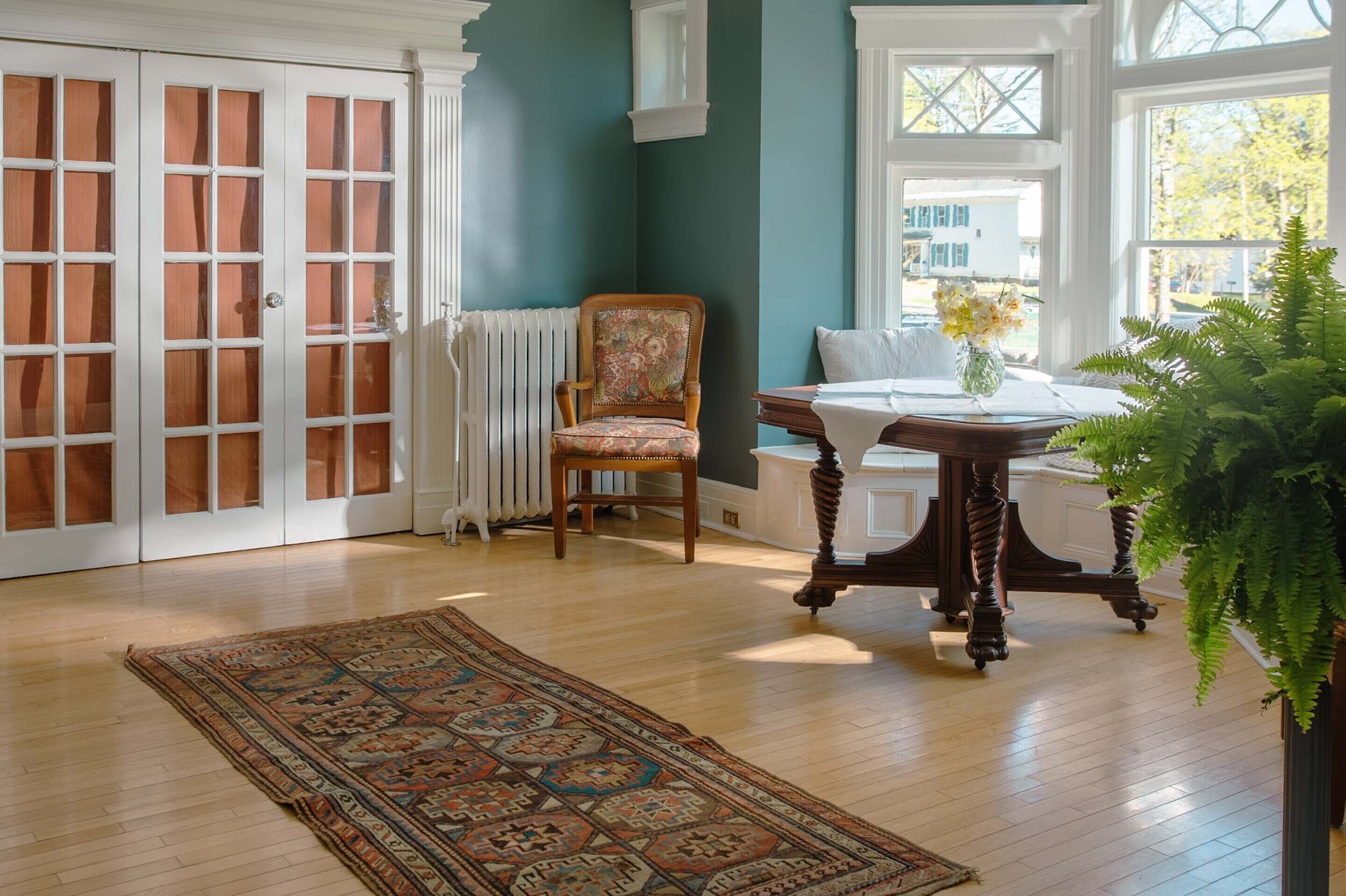 A room with a patterned rug, a wooden chair, a table with a vase, and a large plant.
