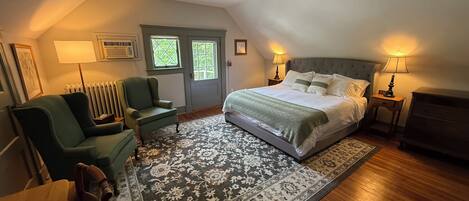 A bedroom with a large bed, two bedside tables with lamps, a green armchair, a wooden dresser, a radiator, and a window with a view of greenery.