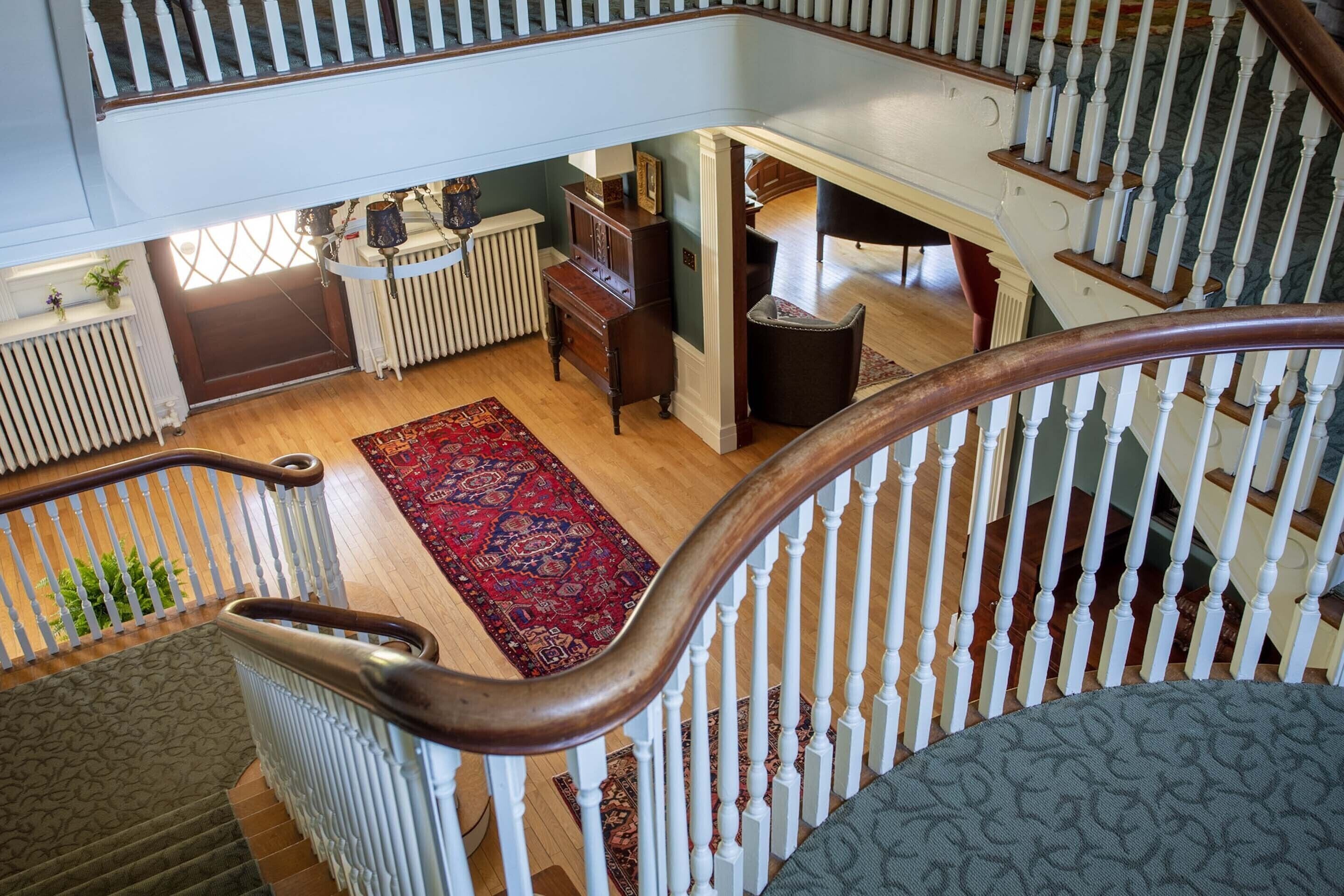 A grand staircase with a wooden handrail and white balusters, leading to a landing with a red patterned rug and a wooden bench.