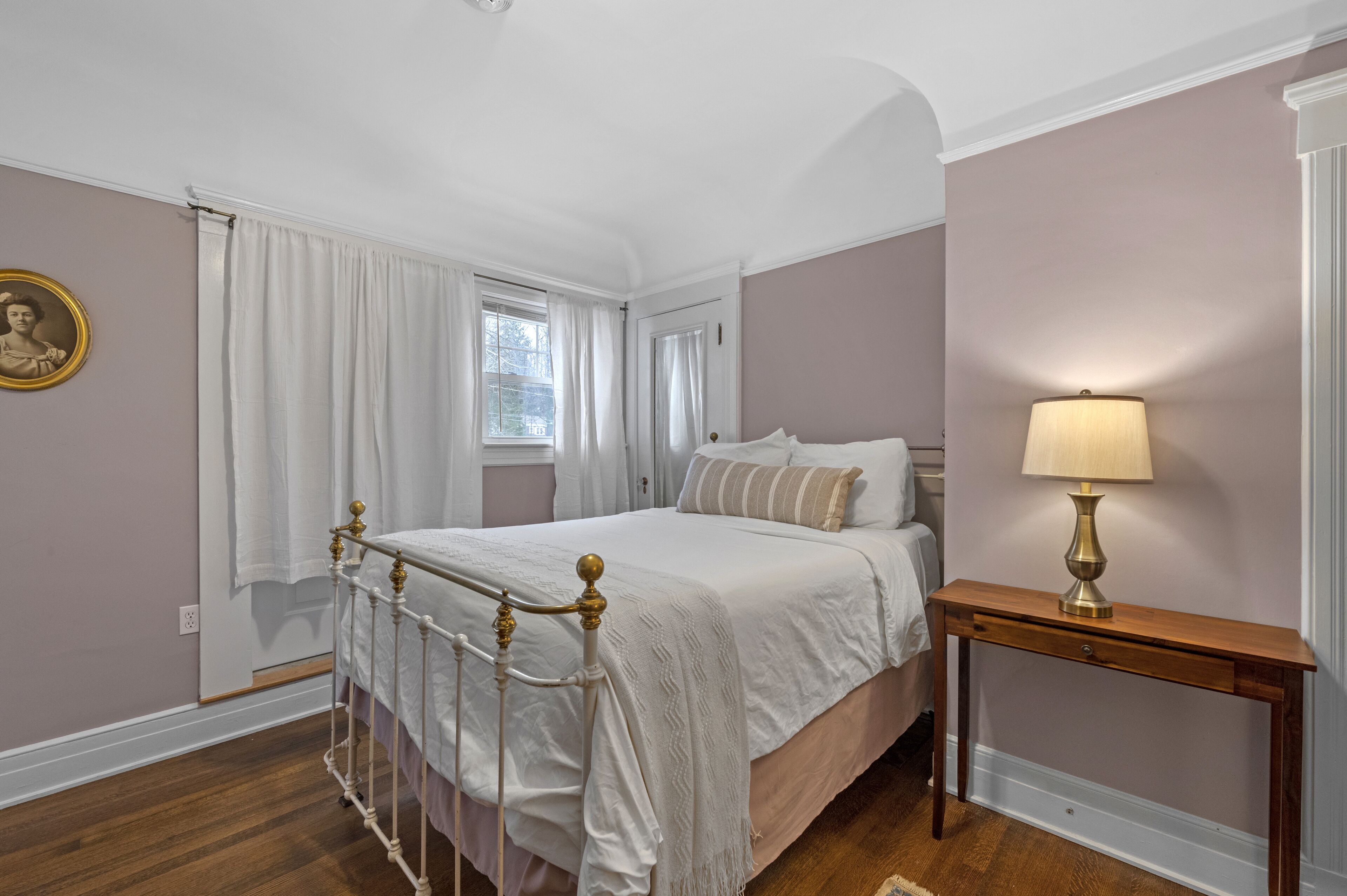 Un dormitorio con cama, una mesita de noche con lámpara, una ventana con cortinas y un cuadro en la pared.